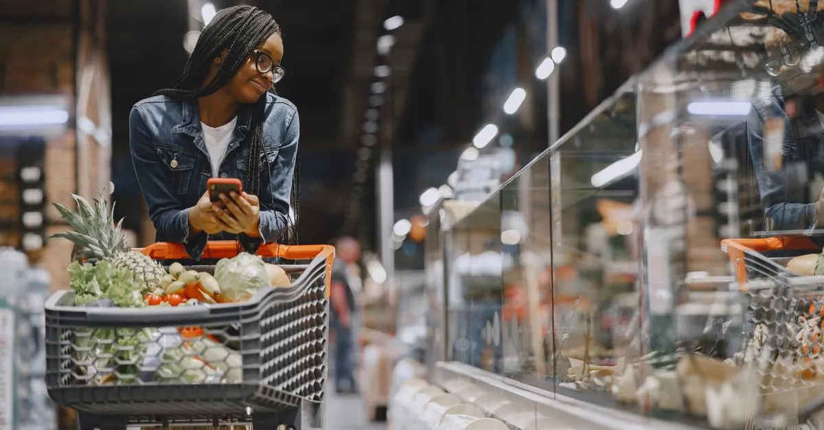 A mom with two young children, one sitting in the shopping cart, looking at a grocery list while navigating an aisle in a well-stocked, budget-friendly grocery store like Aldi. The cart contains a mix of fresh produce and pantry staples.